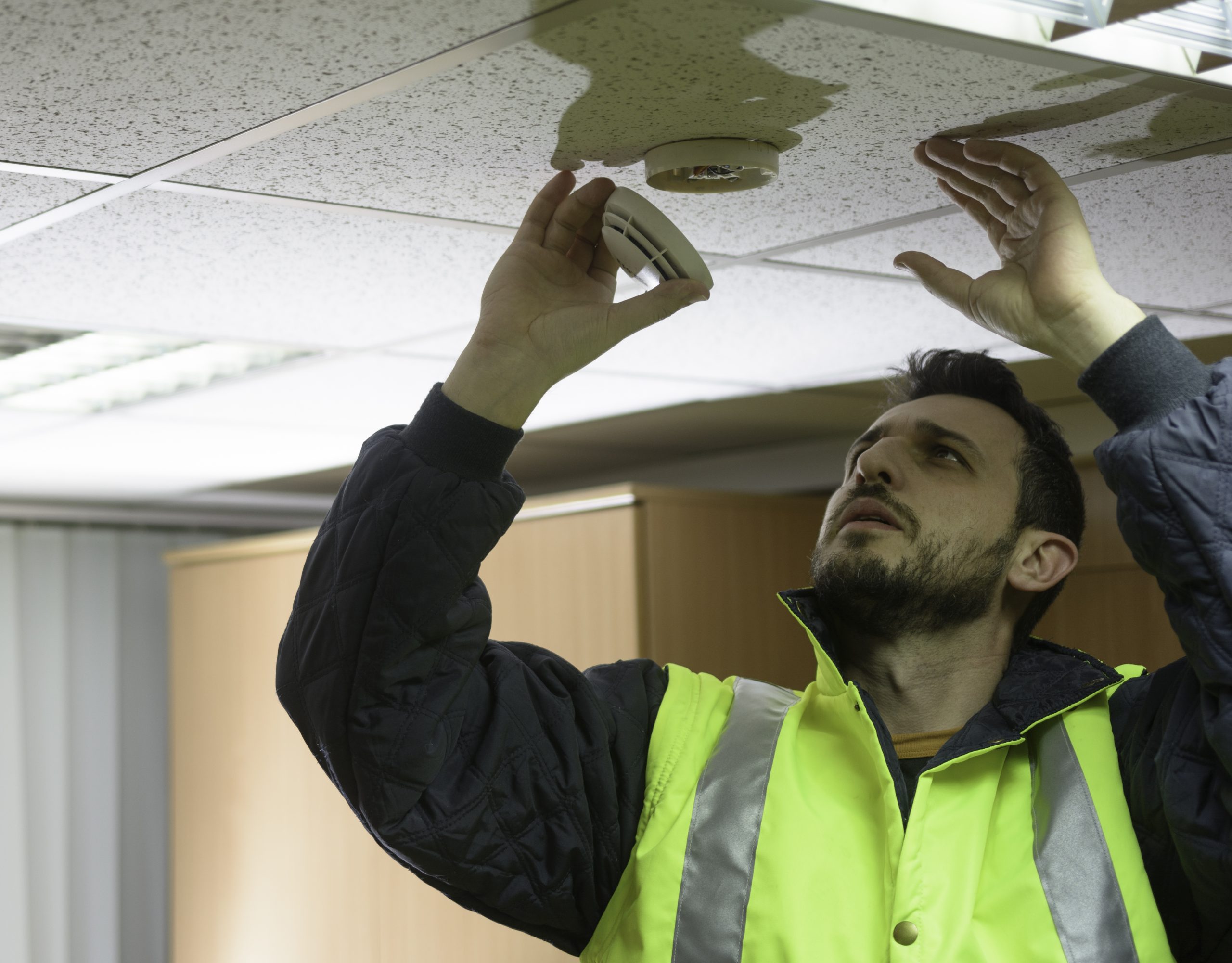 A technician in a bright yellow safety vest examines a fire alarm in the ceiling.